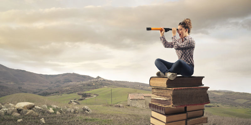 a woman sits atop a stack of books looking thru a spyglass